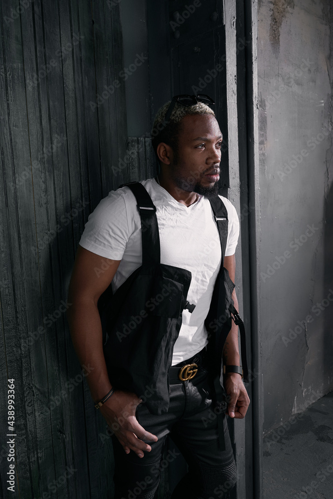 Stylish glamor handsome african man with white short hair wearing white t-shitr, black pants and sunglasses standing against dark wall Stylish glamor handsome african man with white short hair wearing white t-shitr, black pants and sunglasses standing against dark wall