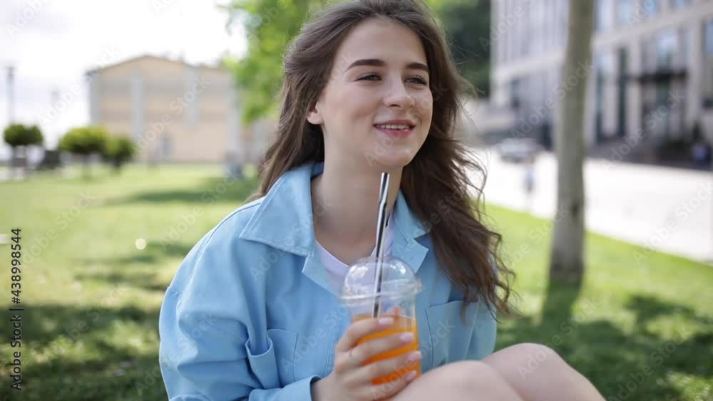 The girl is sitting on the grass with a cocktail. A woman is resting in the city center on a green meadow with a refreshing cocktail. Hot summer day. Happy girl with long hair The girl is sitting on the grass with a cocktail. A woman is resting in the city center on a green meadow with a refreshing cocktail. Hot summer day. Happy girl with long hair