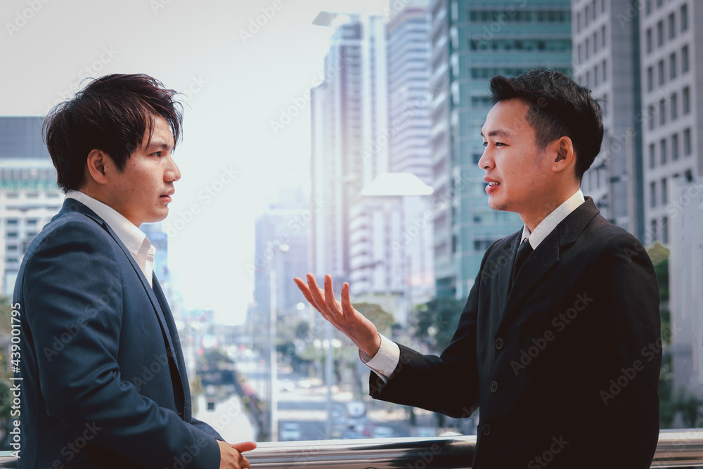 Two successful businessmen in suit talking and discussing at modern city downtown with skyscraper building as blurred background, businesspeople meeting at outside office. Two successful businessmen in suit talking and discussing at modern city downtown with skyscraper building as blurred background, businesspeople meeting at outside office.