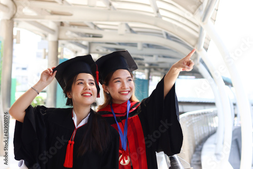 Two happy smiling graduated students, young beautiful Asian women looking at same way, pointing something, so proud on their commencement day, people celebrating successful education on graduation day