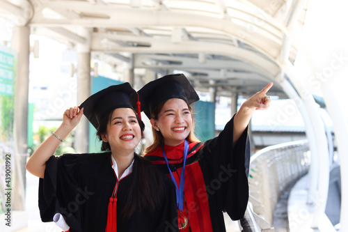 Two happy smiling graduated students, young beautiful Asian women looking at same way, pointing something, so proud on their commencement day, people celebrating successful education on graduation day