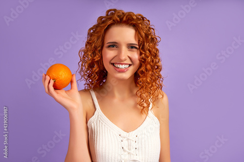 Cute redhead girl with curls holding an orange isolated on a purple background. Proper nutrition. Healthy lifestyle.