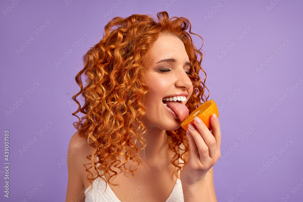 Cute redhead girl with curls eats orange isolated on purple background. Proper nutrition. Healthy lifestyle. Cute redhead girl with curls eats orange isolated on purple background. Proper nutrition. Healthy lifestyle.