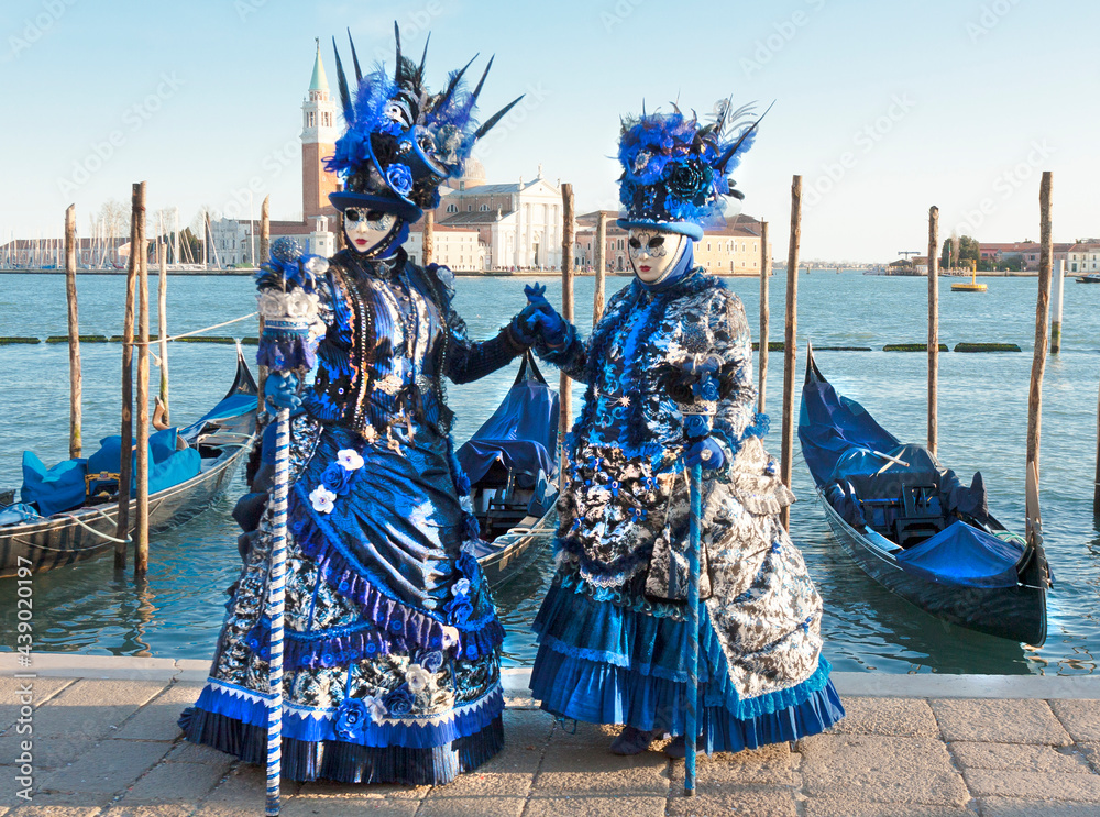 VENICE, ITALY - FEBRAURY 20, 2020: two women in blue carnival constumes and masks on carnival , gondloas and St. George monastery.