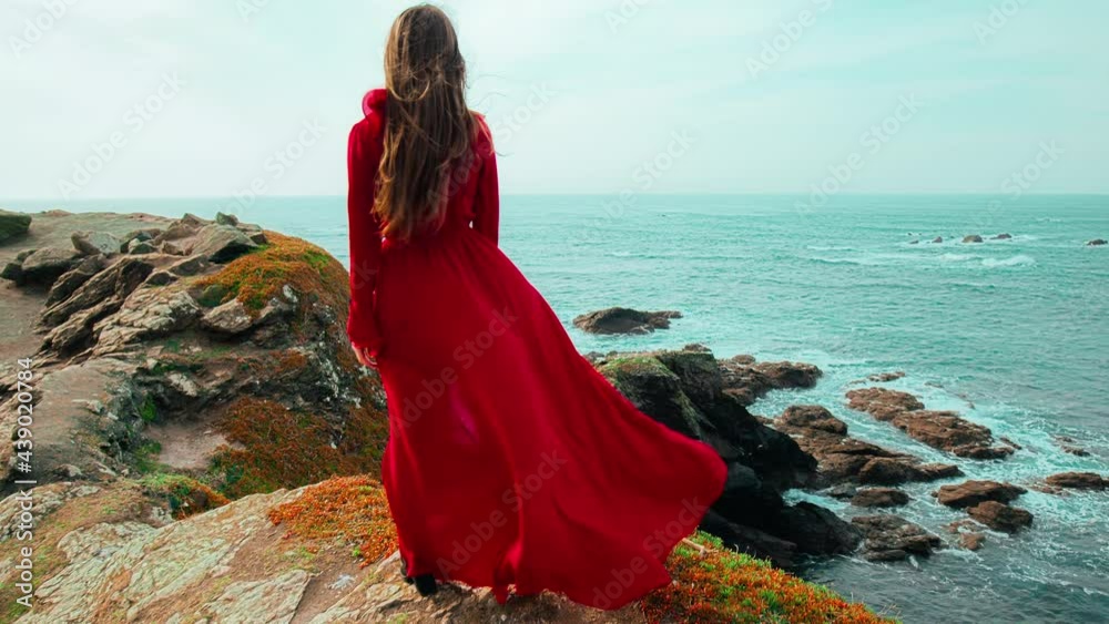 Strong wind blowing a red dress of a young woman standing by the edge of a cliff. A beautiful girl enjoying rocky seashore and scenic landscapes with flying birds in Lizard, Cornwall, England in 4K.
