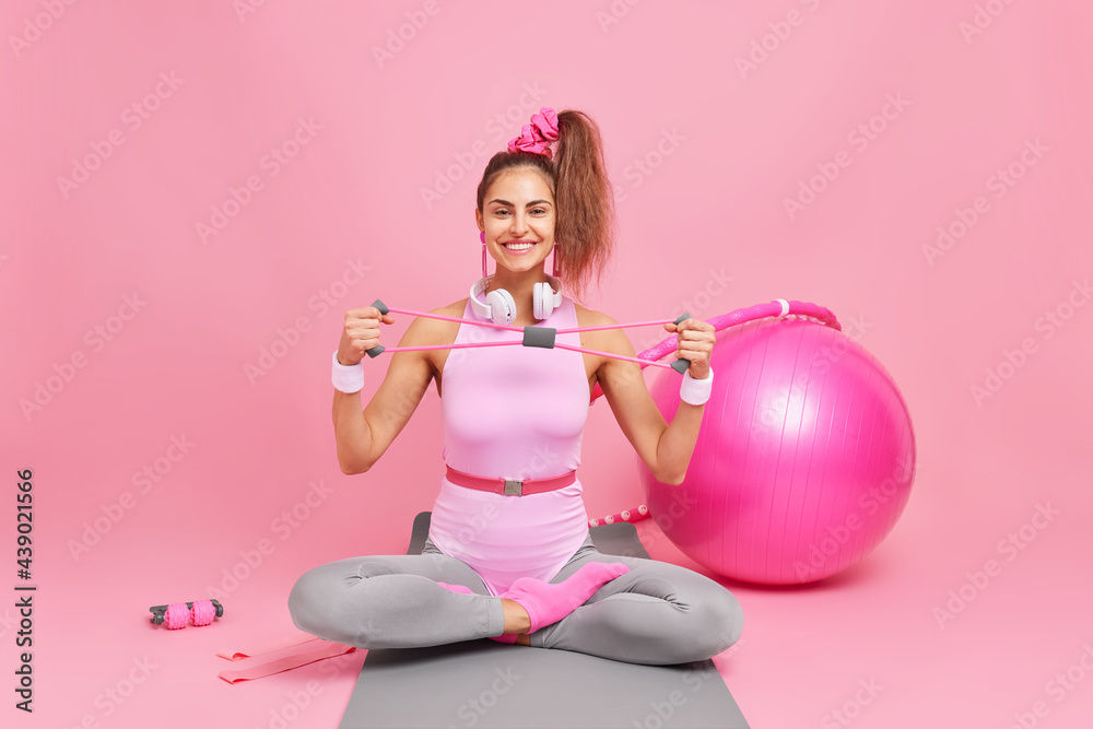Happy fitness female model sits crossed legs on mat stretches expander trains muscles dressed in bodysuit surrounded by swiss ball resistance band hula hoop does exercises for losing weight. Happy fitness female model sits crossed legs on mat stretches expander trains muscles dressed in bodysuit surrounded by swiss ball resistance band hula hoop does exercises for losing weight.