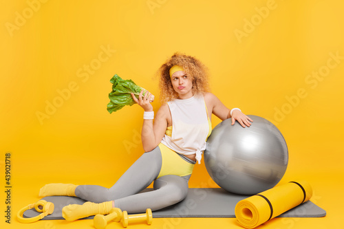 Healthy lifestyle concept. Serious curly haired European woman dressed in activewear keeps to diet holds green vegetable poses on mat with sport equipment isolated over yellow studio background. photo