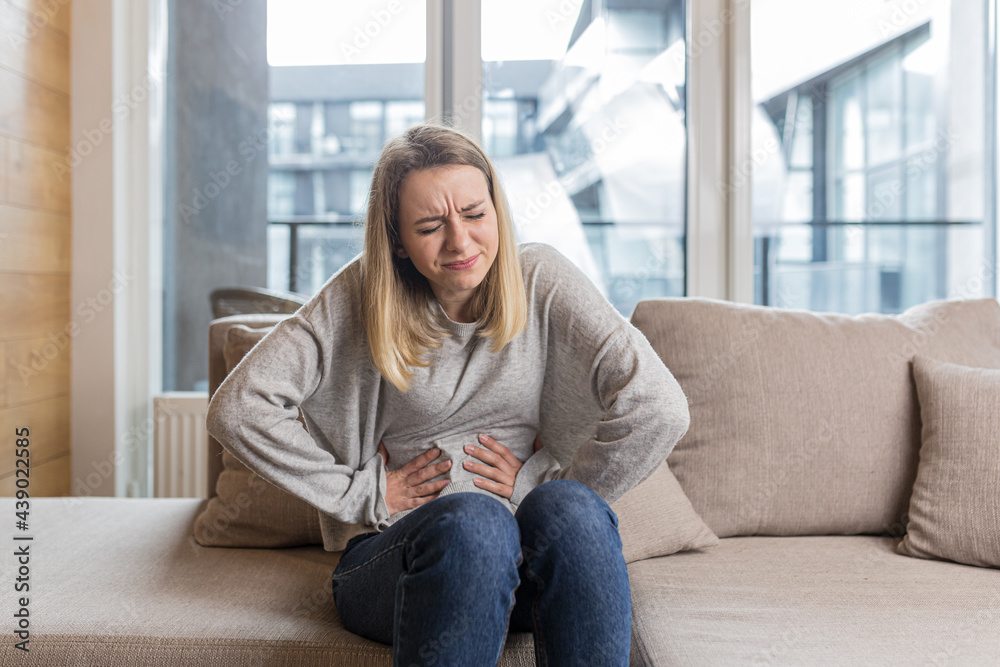 Young woman sitting on sofa holding hands on stomach twisted from pain, poisoned or menstrual pain, woman at home Young woman sitting on sofa holding hands on stomach twisted from pain, poisoned or menstrual pain, woman at home