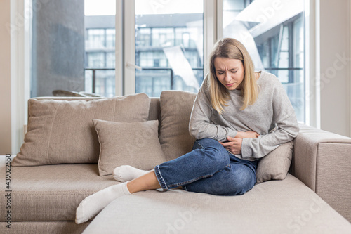 Young woman sitting on sofa holding hands on stomach twisted from pain, poisoned or menstrual pain, woman at home