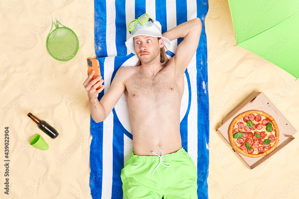 Surprised topless man stares at smartphone display lies on striped blue towel wears sun hat green shorts sunbathes during day off has recreation outdoor eats pizza drinks beer has lazy time.