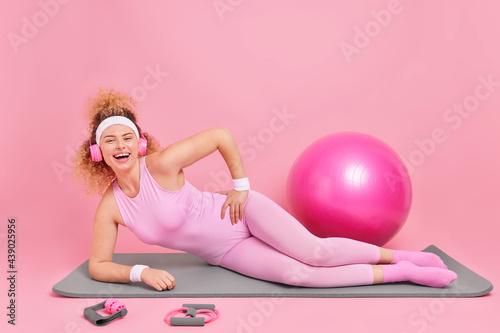 Positive slim curly haired young woman stands in plank pose on fitness mat dressed in activewear listens music via headphones uses sport equipment isolated over pink background. Workout at home photo