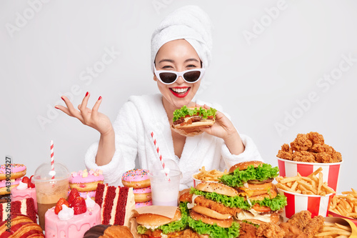 Indoor shot of happy Asian woman raises hand smiles sincerely holds tasty hamburger wears sunglasses dressed in domestic clothes affords herself eating junk food isolated over white studio wall