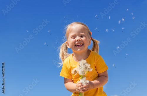A little girl with a bunch of white dandelions against blue sky. Smiling child in a yellow t-shirt, summer holidays.