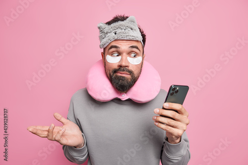 Indoor shot of hesitant puzzled man shrugs shoulders holds mobile phone checks newsfeed wears blindfold neck pillow patches under eyes isolated over pink background surprised to read shocking news © wayhome.studio 
