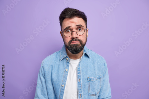 Handsome frustrated bearded man with pity expression frows face looks unhappily being bothered faces troubles wears round spectacles denim shirt isolated over purple background. Negative emotions photo