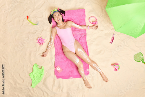Carefree smiling Asian teenage girl with perfect figure wears sunbathing suit poses on soft pink towel applies sunscreen on body while sunbathing spends leisure time at tropical sandy coastline