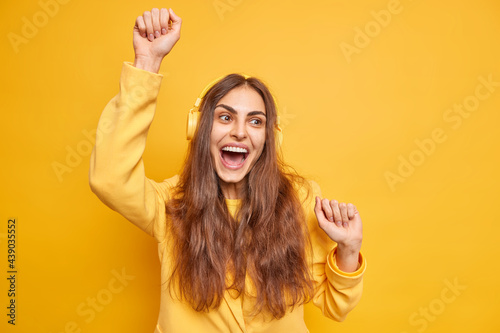 Positive dark haired young woman dances carefree keeps arms raised up enjoys favorite music keeps mouth opened poses against vivid yellow background. Upbeat European female moves rhythmically
