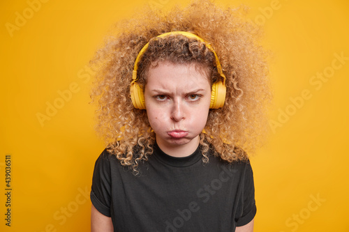 Portrait of young sulking young woman with curly hair blows cheeks looks angrily being offended listens music via headphones dressed in black t shirt isolated over yellow background has bad mood