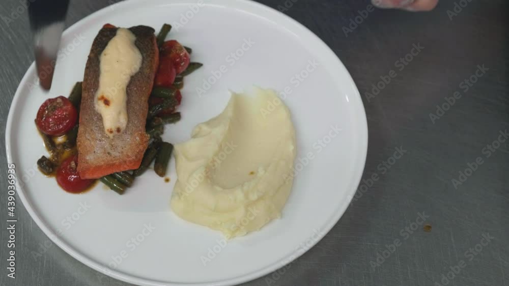The chef's hands place the fried red fish fillets on the fried vegetables in a plate of mashed potatoes. Then he burns the cheese on the fish with a burner. Restaurant serving The chef's hands place the fried red fish fillets on the fried vegetables in a plate of mashed potatoes. Then he burns the cheese on the fish with a burner. Restaurant serving
