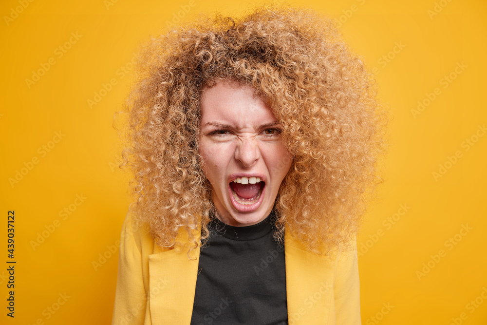 Portrait of outraged woman with curly bushy hair screams angrily has quarrel expresses negative emotions wears formal clothing isolated over yellow background. Aggressive annoyed female model Portrait of outraged woman with curly bushy hair screams angrily has quarrel expresses negative emotions wears formal clothing isolated over yellow background. Aggressive annoyed female model