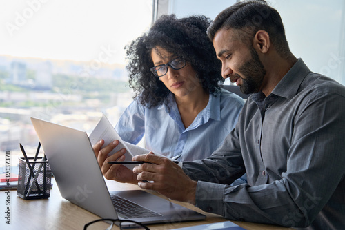 Multiethnic male indian mentor and female African American intern sitting at desk with laptop doing paperwork together discussing project financial report. Corporate business collaboration concept. photo