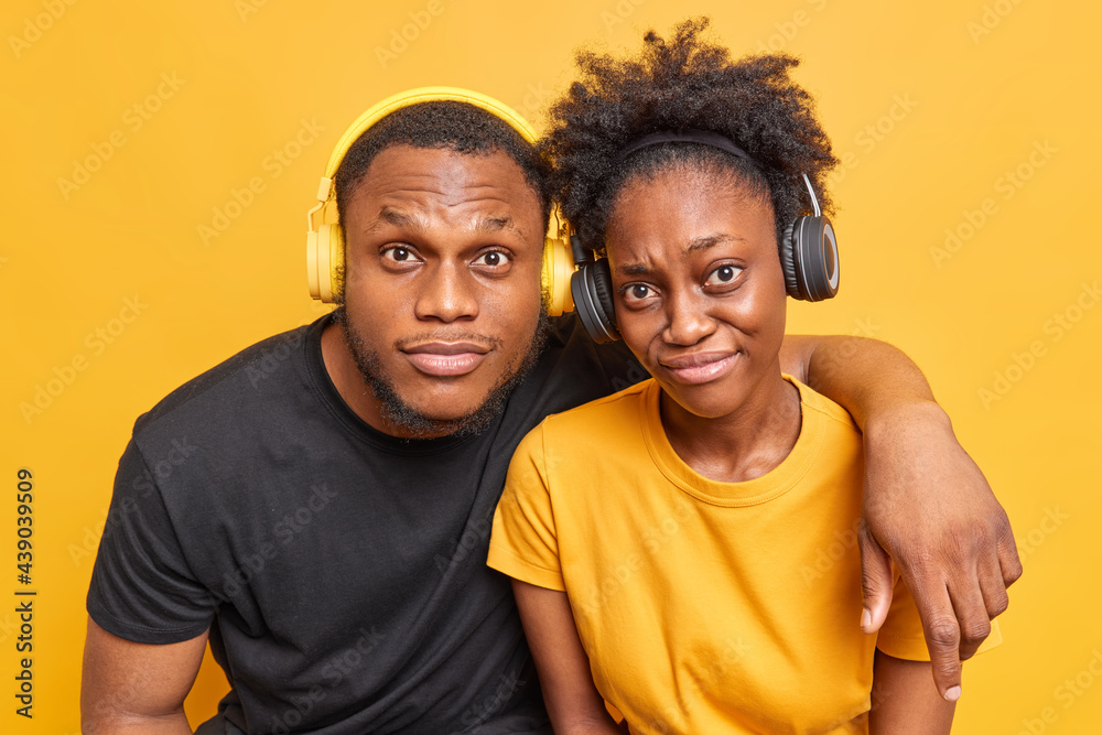 Fototapeta premium Photo of friendly dark skinned woman and man embrace and look attentively at camera dressed casually listen music via headphones isolated over vivid yellow background learn foreign languages together
