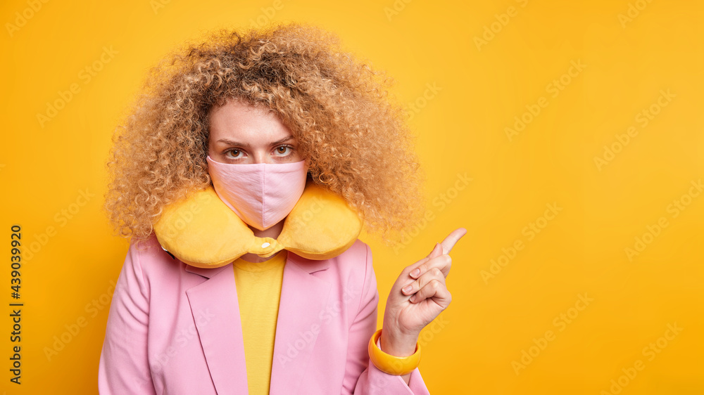 Studio shot of beautiful curly haired young woman looks seriously at camera wears disposable face mask as protection against virus indicates away on copy space isolated over yellow background Studio shot of beautiful curly haired young woman looks seriously at camera wears disposable face mask as protection against virus indicates away on copy space isolated over yellow background
