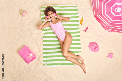 Top view of smiling relaxed curly haired woman with perfct figure lies on striped towel at beach listens music via headphones takes sunbath enjoys sun rests near sea dressed in bathing suit. photo