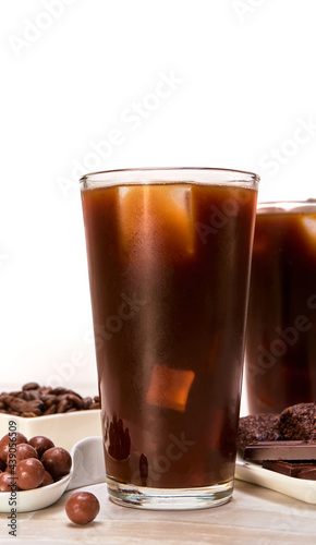 american iced coffee accompanied by chocolate pearls, vegan brownies and a couple of chocolate bars on a marble surface and a white background. copy space for advertising. luxury concept.