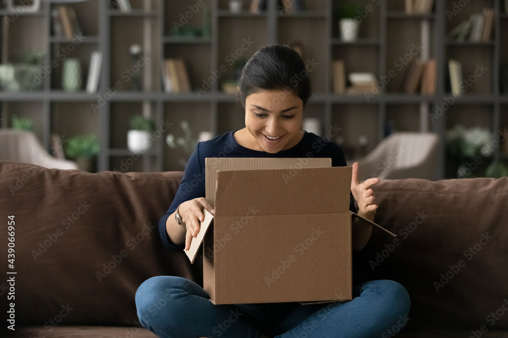What a nice surprise. Joyful teenage indian female addressee sit on sofa looking inside big cardboard box received by mail service. Happy interested young woman opening carton postal parcel enjoy gift - obrazy, fototapety, plakaty 