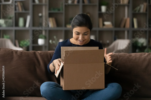 What a nice surprise. Joyful teenage indian female addressee sit on sofa looking inside big cardboard box received by mail service. Happy interested young woman opening carton postal parcel enjoy gift photo