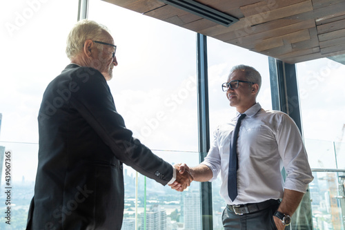 Two business men discussing their business together in a lobby of a hotel. They both wearing white shirt. The older one also wear black jacket. The scene outside is cityscape. They are shaking hand.