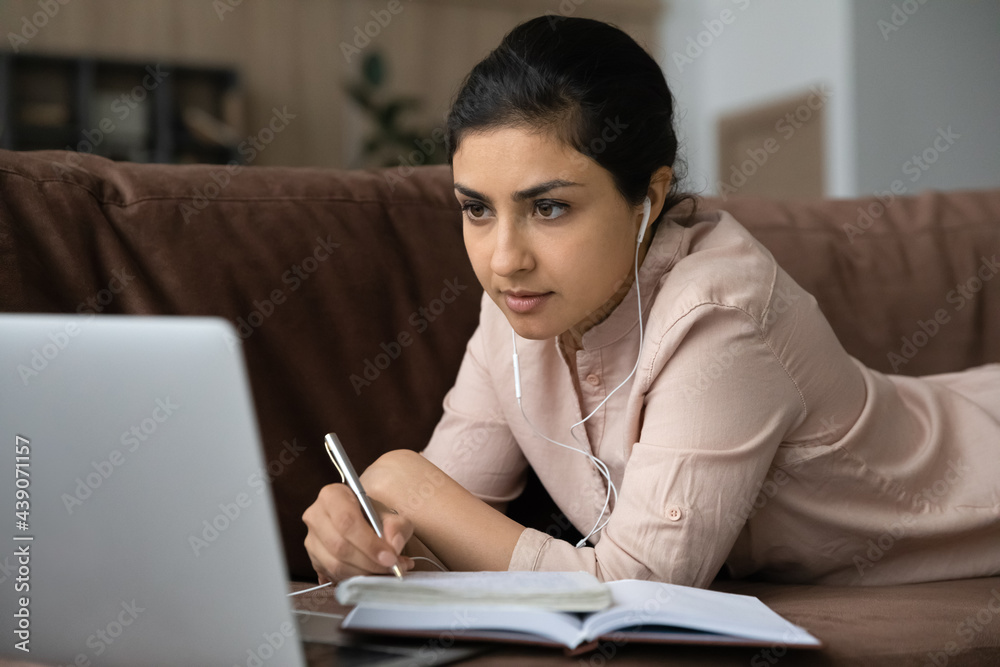 Focused indian female student in wired headphones lie on sofa in comfy pose before laptop screen write checkup test exam in paper copybook. Young lady study business course at web platform take notes Focused indian female student in wired headphones lie on sofa in comfy pose before laptop screen write checkup test exam in paper copybook. Young lady study business course at web platform take notes