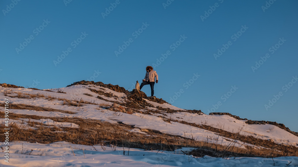 Dry grass and snow on the hillside are illuminated by the rising sun. A man in a down jacket and a hood is standing against the background of the blue sky. Nearby is a pyramid of stones. Siberia