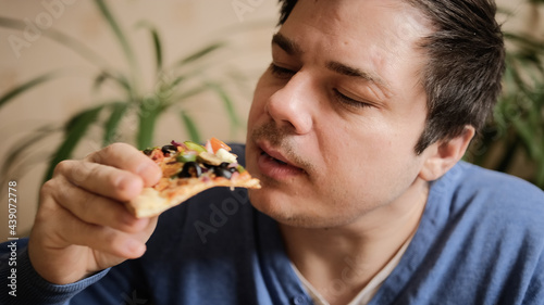 A slightly shaved young man eats a vegetarian pizza with mushrooms, tomatoes, peppers and cheese. © chekart