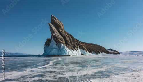 A bizarre rocky island rises above a frozen lake against a blue sky. Sharp peak, steep slopes devoid of vegetation. The base is covered with icicles. Cracks in the ice. Baikal