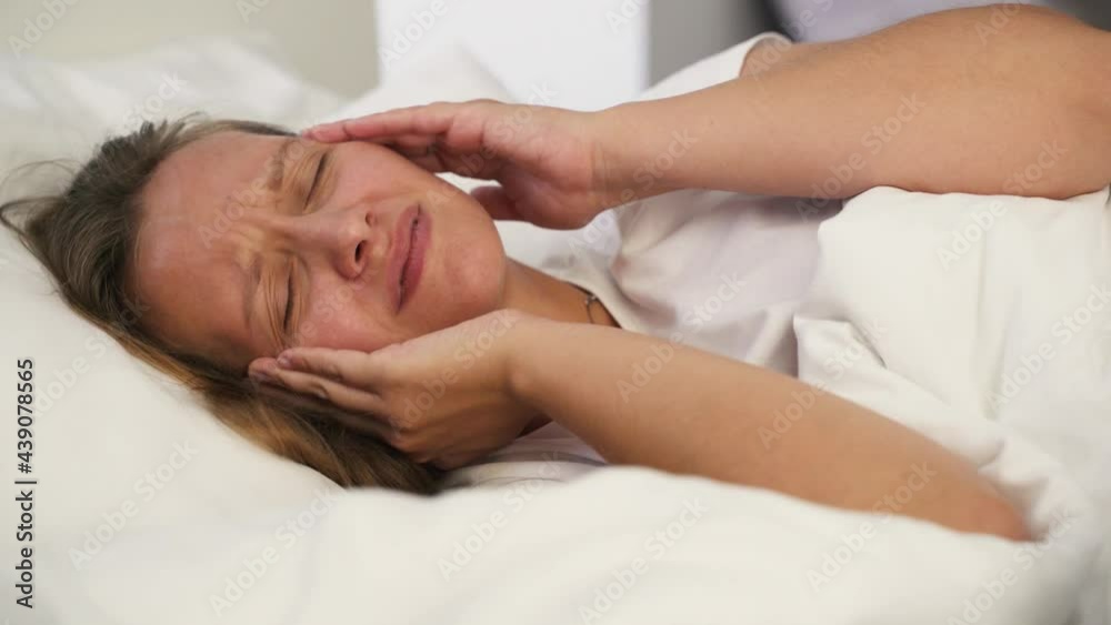Young irritated woman cannot sleep from headache. Lies alone in bed in the bedroom under white blanket on pillow in white pajamas and massages her temples for migraines. Sleep disorder, insomnia Young irritated woman cannot sleep from headache. Lies alone in bed in the bedroom under white blanket on pillow in white pajamas and massages her temples for migraines. Sleep disorder, insomnia