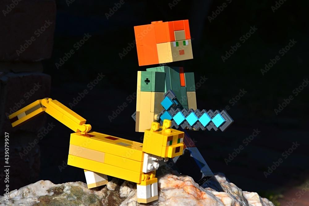 Naklejka premium LEGO Minecraft large action figure of Alex with diamond sword standing on rock with friendly Ocelot cat, dark background.