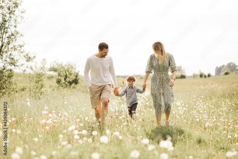 Fototapeta premium Cheerful parents having fun with their little son 3 years in summer field while holding hands and walking together.