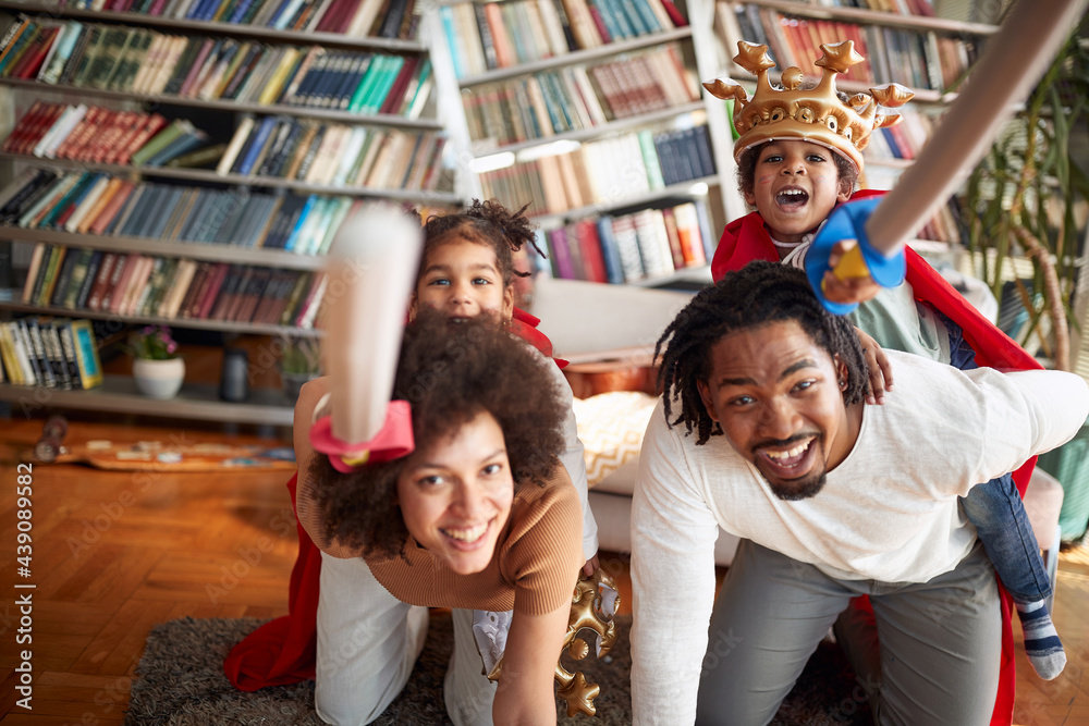 Fototapeta premium Cheerful kids are shouting and posing while riding their parents in a knights game at home. Family, home, love, together