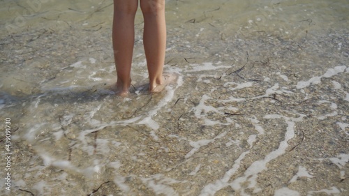 Legs of a child jumping over the waves on the seashore. Algae float on the water on the beach. Summer vacation at sea.