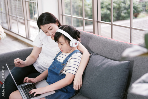 Smiling Asian mother and little girl child is relaxing with headphone and laptop for listening to online music or cartoon movie in social media on sofa in living room. Education and technology concept