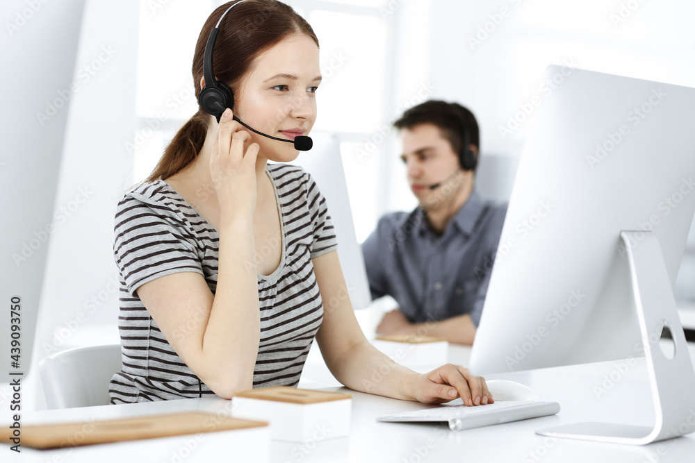Casual dressed young woman using headset and computer while talking with customers online. Group of operators at work. Call center, business concept Casual dressed young woman using headset and computer while talking with customers online. Group of operators at work. Call center, business concept