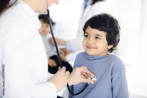 Doctor-woman examining a child patient by stethoscope. Cute arab boy and his brother at physician appointment. Medicine concept