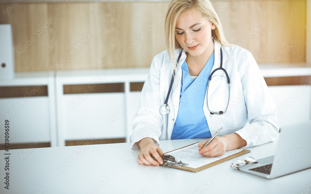 Woman-doctor at work while sitting at the desk in sunny clinic. Blonde cheerful physician filling up medication history record form Woman-doctor at work while sitting at the desk in sunny clinic. Blonde cheerful physician filling up medication history record form