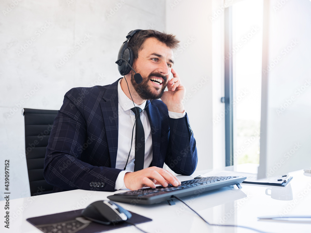Cheerful man in suit sitting in office, wearing headphones, typing and smiling. Cheerful man in suit sitting in office, wearing headphones, typing and smiling.