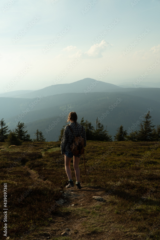 Fototapeta premium Woman on the summit of Králický Sněžník between Poland and the Czech Republic, at sunset, high mountain meadow, with Czarna Gora in the background. Classic hiking with a backpack.