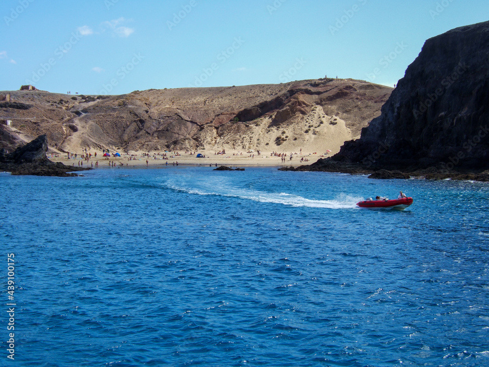 View of a bay with a big sandy beach and many bathers between some rocks with deep blue sea on which a small red rubber dinghy is sailing in Lanzarote