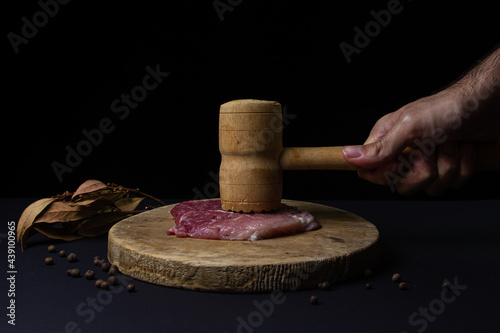 A man's hand beats the meat with a kitchen hammer. Wooden kitchen hammer for meat. Meat on black background