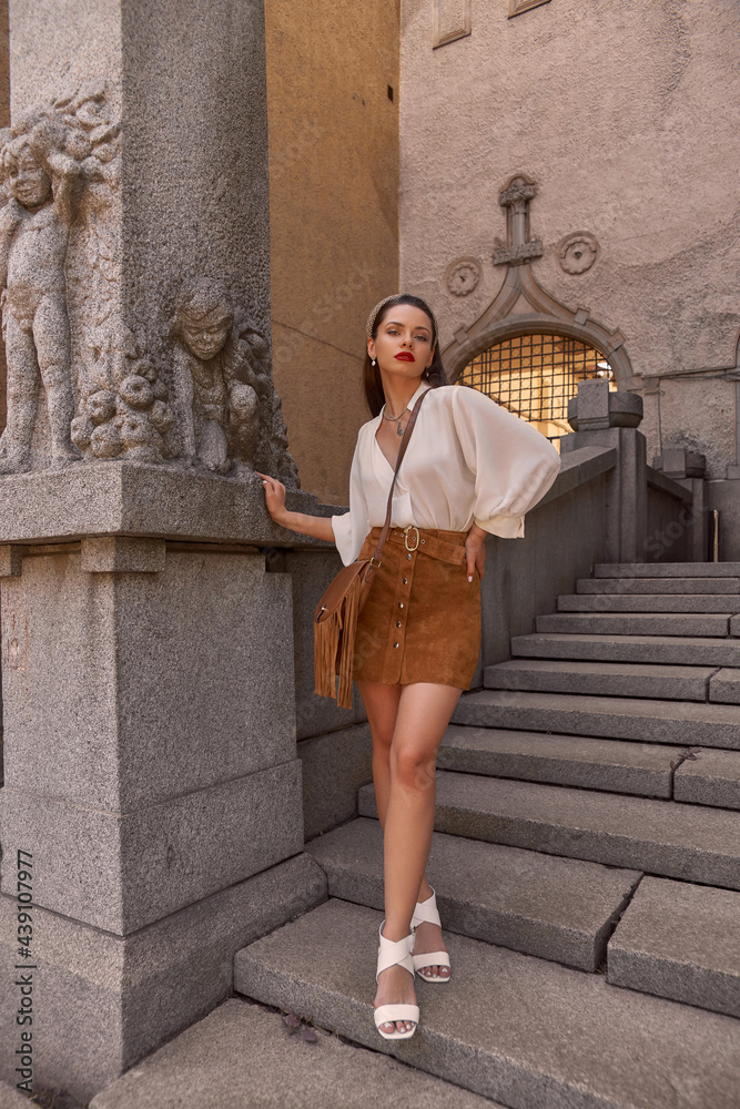 Full length outdoor portrait of young beautiful woman in white blouse, brown skirt, in white shoes, with handbag standing and posing at stone stairs